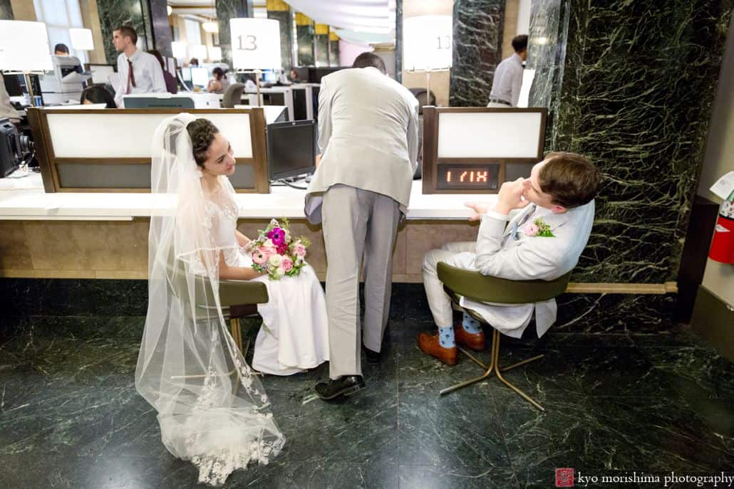 Bride and groom lean back in chairs to smile at one another as witness signs their wedding certificate during NYC City Hall wedding. Bouquet in bride's lap. Long veil pooled on floor. Summer NYC elopement wedding. Homecoming florist.