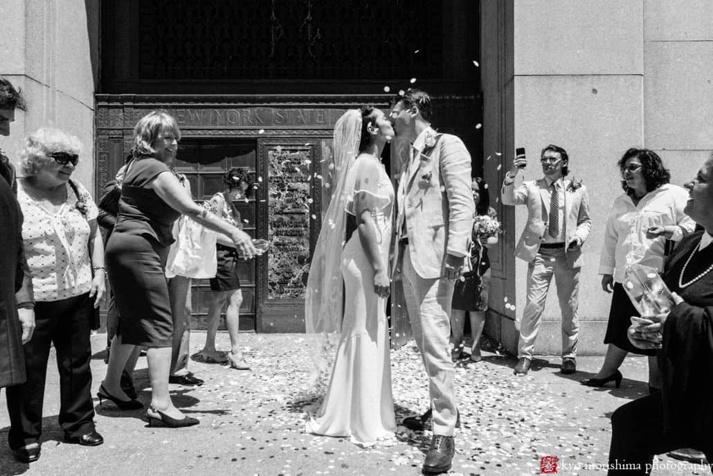 bride and groom kiss outside City Hall as guests throw petals. NYC City Hall elopement photographer. Black and white wedding photography. Summer NYC wedding. 