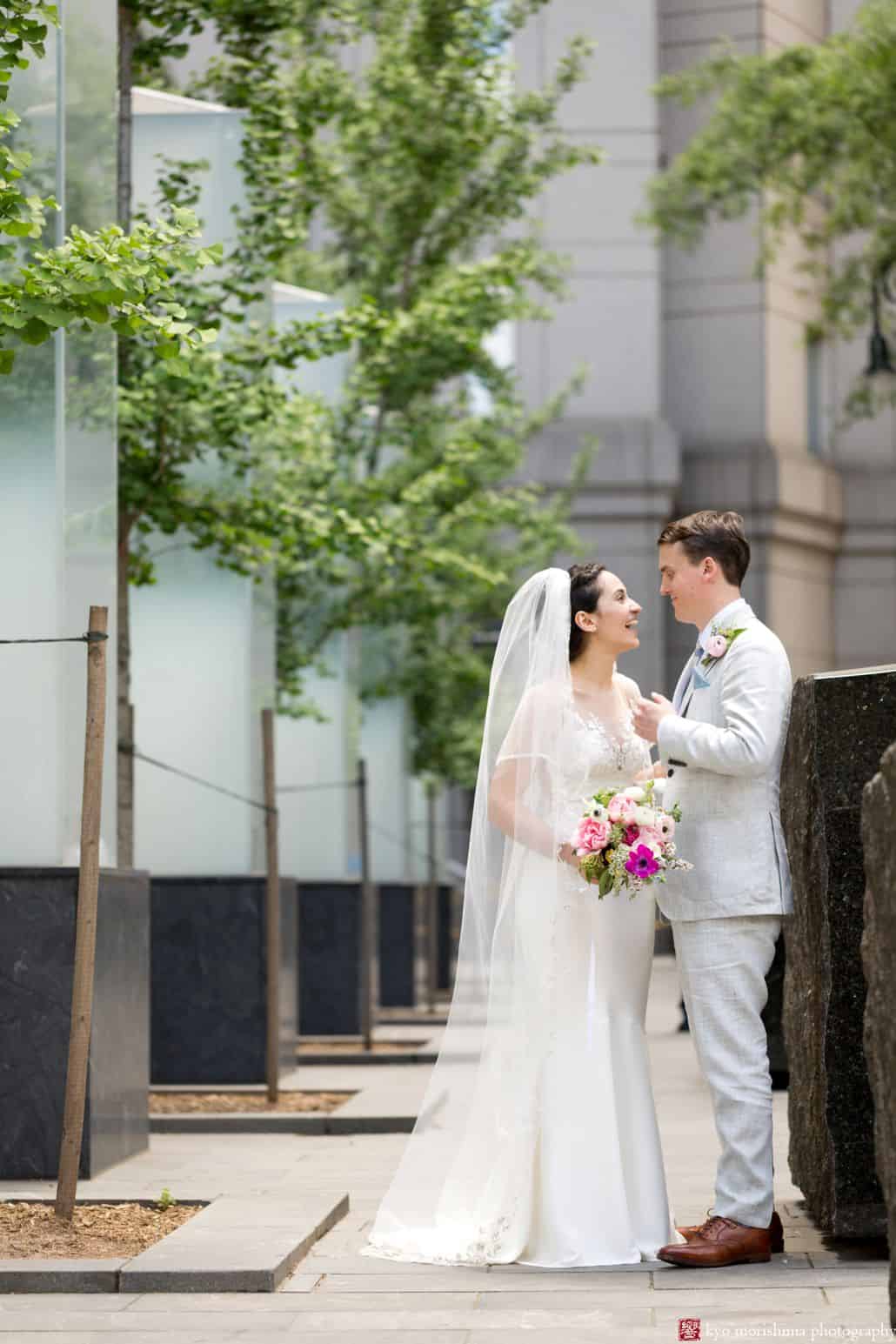 NYC City Hall wedding photos: Bride and groom smile at one another outside City Hall. Pink, fuschia and white bouquet, groom wears light gray linen suit. Small trees in the background. Summer NYC City Hall wedding photos. Homecoming florist. 