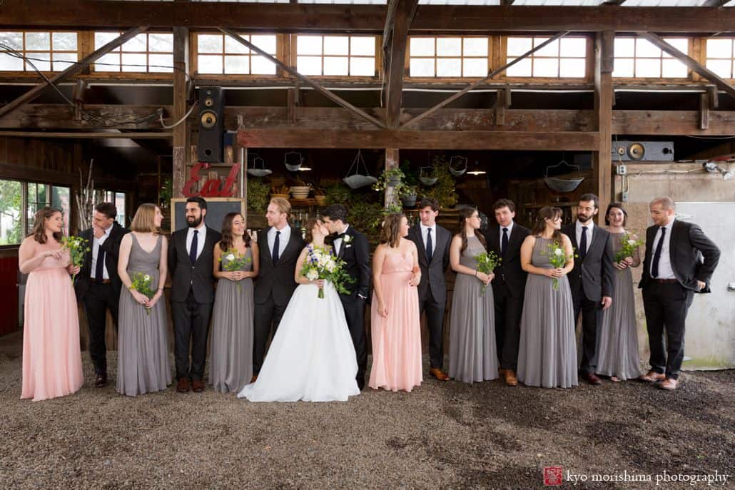 Bride and groom kiss surrounded by wedding party. Large wedding party. gray and peach bridesmaid dresses, charcoal gray groomsmen suits. Designer Loft wedding gown, Fleur di re wedding bouquets. Blooming Hill Farm wedding photographer. Hudson Valley Kyo Morishima Photography