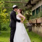 bride and groom kiss outside at Blooming Hill Farm wedding. Stone path, grass, greenery backdrop, rustic wooden window boxes, wooden branch archway, Designer Loft bridal gown, New York Jewish wedding photographer. Summer wedding. groom wears bow tie and kippah.