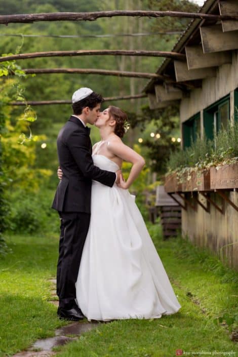 bride and groom kiss outside at Blooming Hill Farm wedding. Stone path, grass, greenery backdrop, rustic wooden window boxes, wooden branch archway, Designer Loft bridal gown, New York Jewish wedding photographer. Summer wedding. groom wears bow tie and kippah.