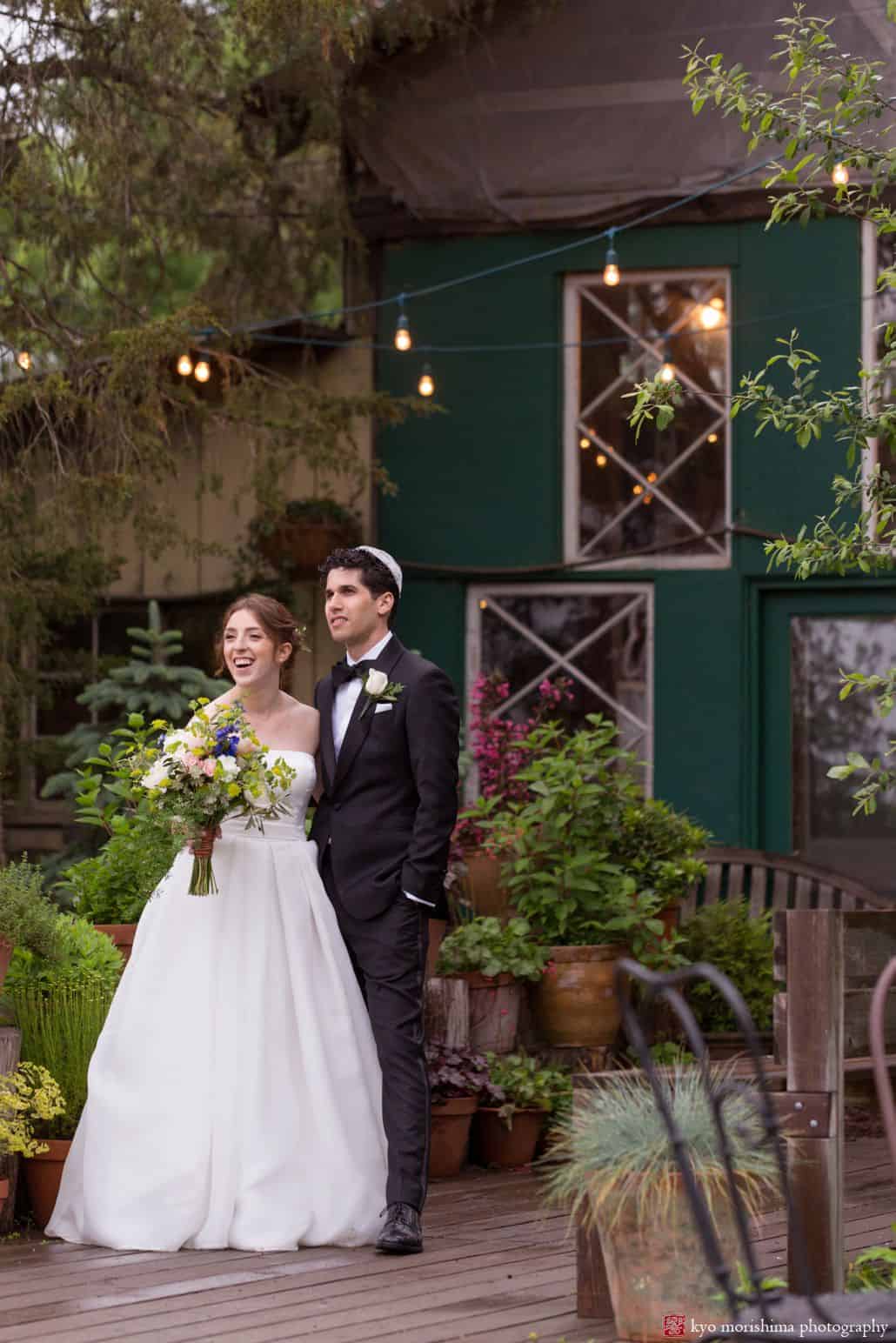 Bride and groom stand together and look on at Blooming Hill Farm wedding. Fleur di re bridal bouquet, Designer Loft wedding dress, green rustic cottage, greenery and potted plants on wooden deck, overhead string lights, NYC Jewish Summer wedding photographer. groom wears bow tie and kippah Hudson Valley Kyo Morishima Photography