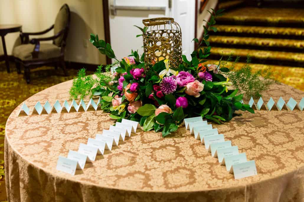 wedding place card table, gold damask tablecloth, gold lantern centerpiece surrounded by fuscia and peach flowers with green leafy fillers, roses, peony, glossy leaves, Kristin Rockhill florist, Nassau Inn, Princeton NJ wedding photographer.