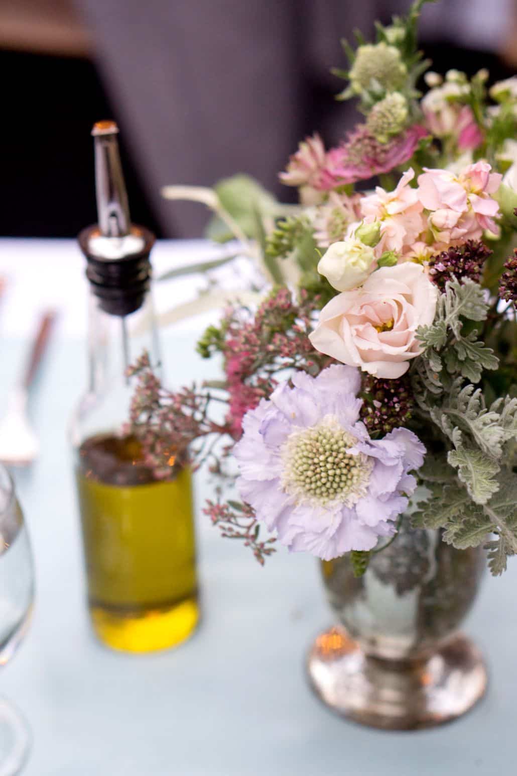 wedding table centerpiece next to bottle of olive oil, vintage colors, white, pink, dusty rose, silver leaves in a silver vase, Opalia florist, Frankie's 457 Spuntino, Brooklyn, NY wedding photographer.