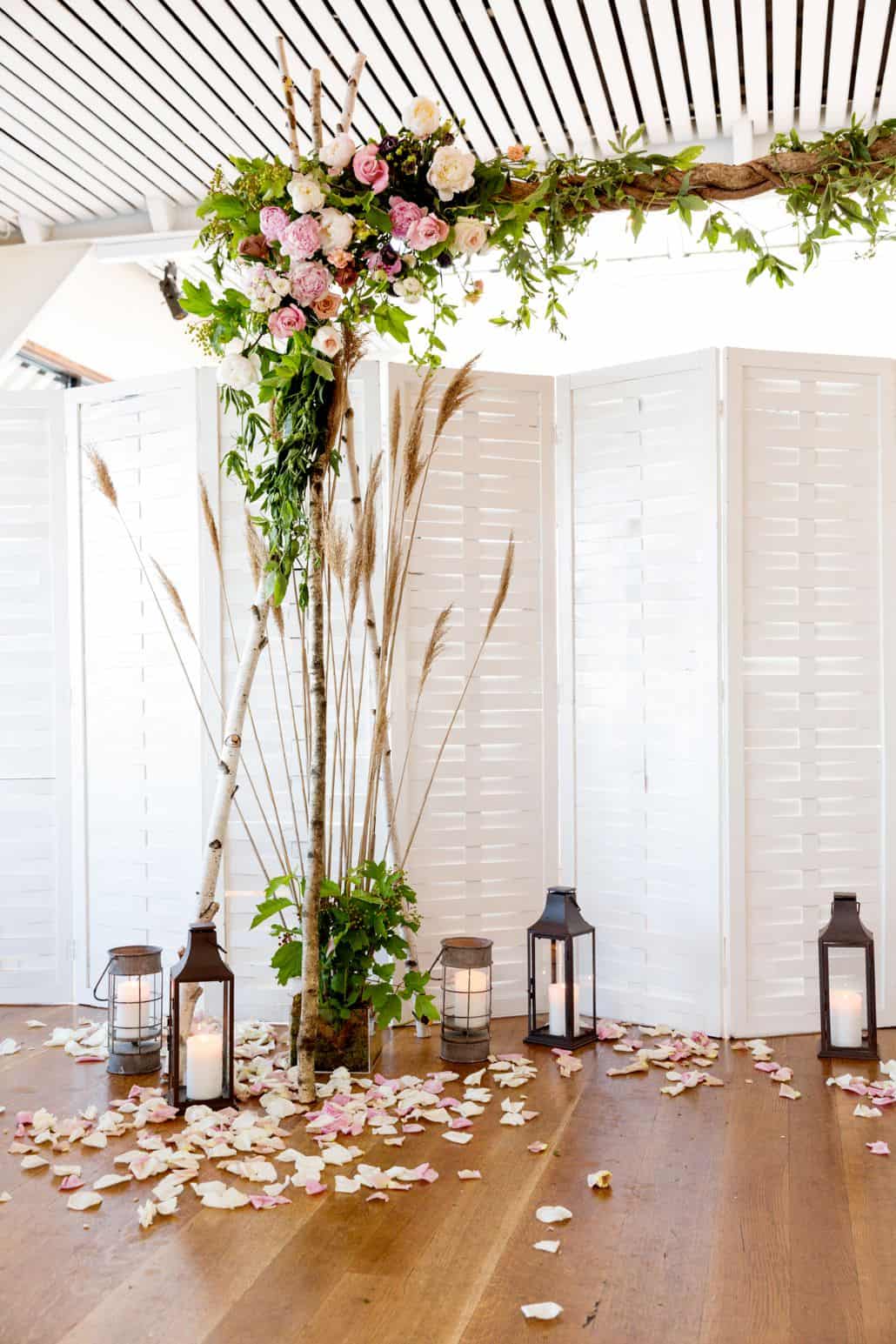 white rose petals and lanterns scattered on wooden floor under floral archway, white shutter floor screen, white slatted ceiling, Quatre Coeur florist, The water Club, NYC wedding photographer.