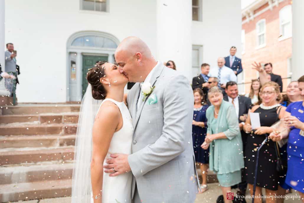 Bride and groom kiss on steps outside Hunterdon County courthouse as guests throw confetti, BHLDN wedding dress, men's warehouse suit, intimate Summer New Hope, PA wedding photographer.