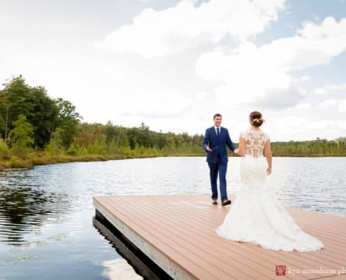 Bride and groom stand at end of dock overlooking a pond and treeline at Woodloch Pines in the Poconos, Castle Couture wedding gown, lace back wedding dress, navy groom's suit, outdoor wedding photographer, resort wedding.