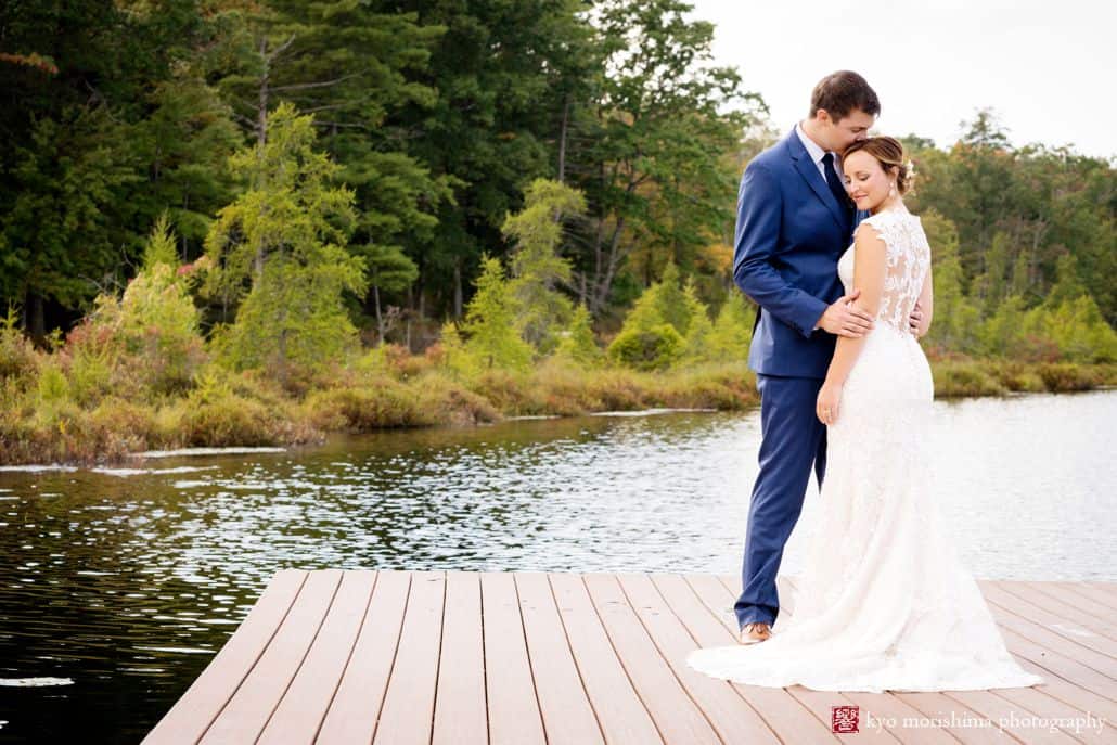 Groom in navy suit kisses bride's head at end of dock overlooking pond and forest at Woodloch Pines resort, Castle Couture wedding gown, outdoor poconos wedding photographer.