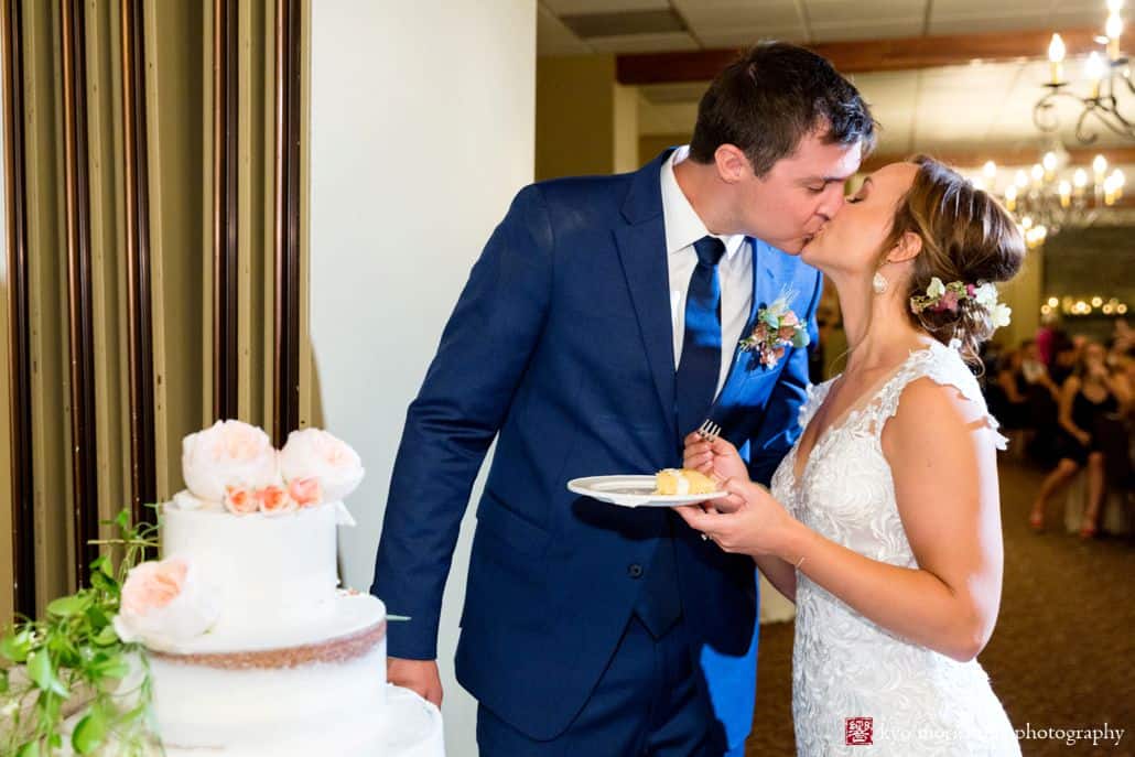 Bride and groom kiss next to wedding cake while bride holds plate with slice of cake, Castle Couture wedding gown, cobalt blue groom's suit, Fox Hill Farm Experience boutineer and bridal hair flowers, burgundy and white flowers with eucalyptus, naked wedding cake with peonies, Woodloch Pines reception, Poconos resort wedding photographer.