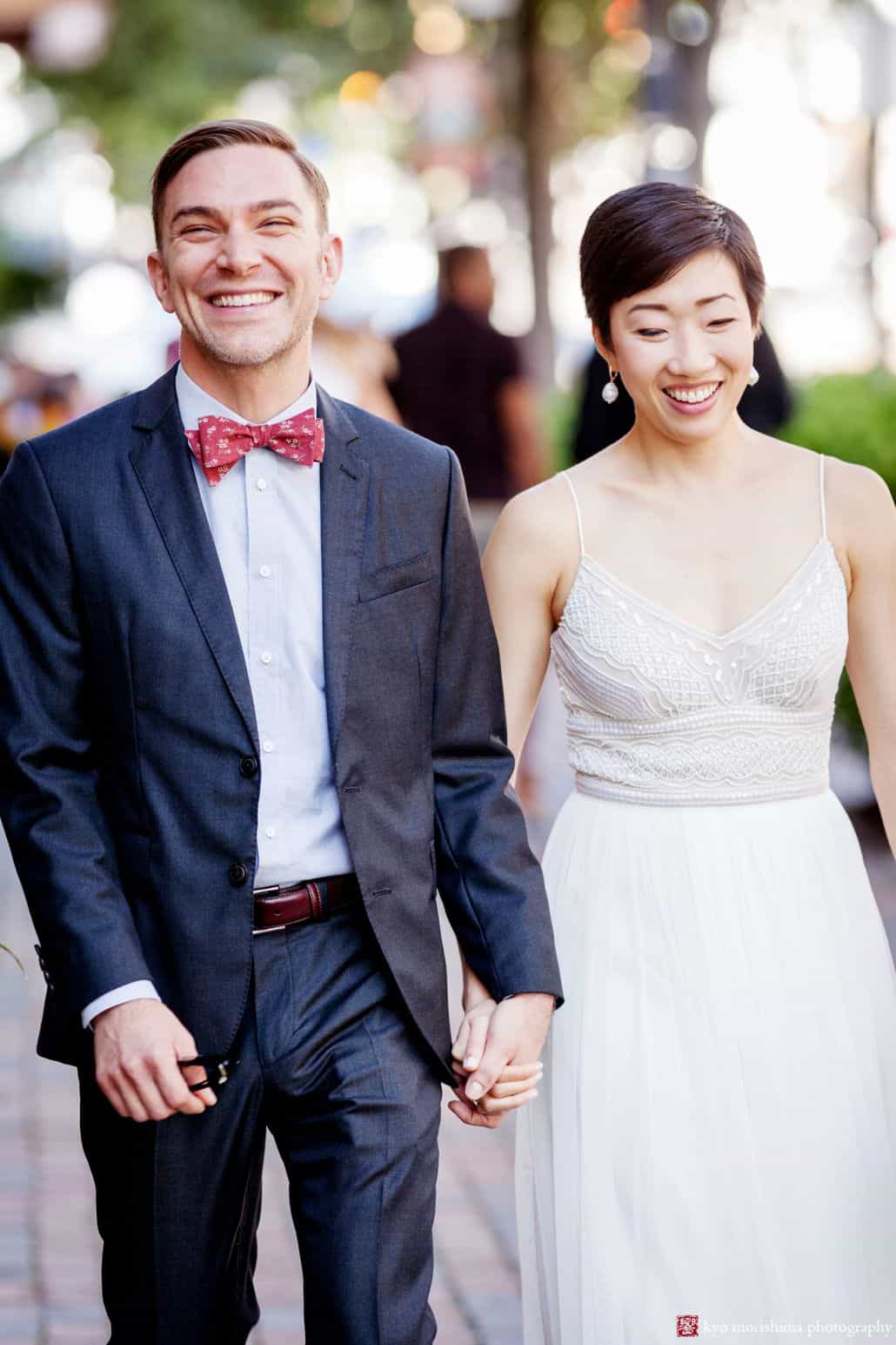 Candid wedding portrait of bicultural couple walking down city street on September wedding day; groom wears red bow tie