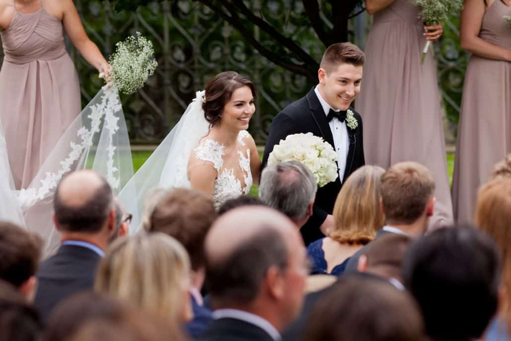 Bride and groom, seen over the tops of their guests' heads, depart the wedding ceremony at Jasna Polana. Bride wears Pronovias. Flowers by Janet Makrancy.