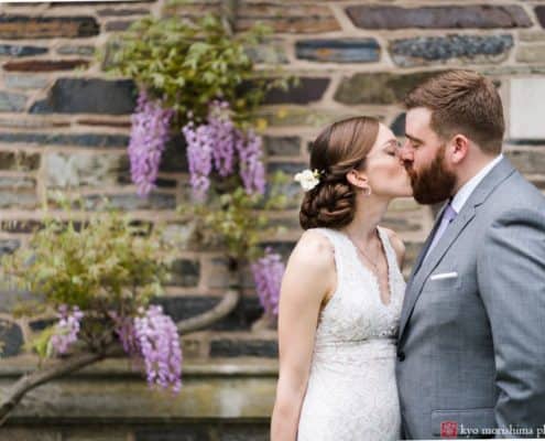 Bride and groom kiss in front of stone wall with wisteria blooms during May wedding in Princeton