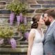 Bride and groom kiss in front of stone wall with wisteria blooms during May wedding in Princeton