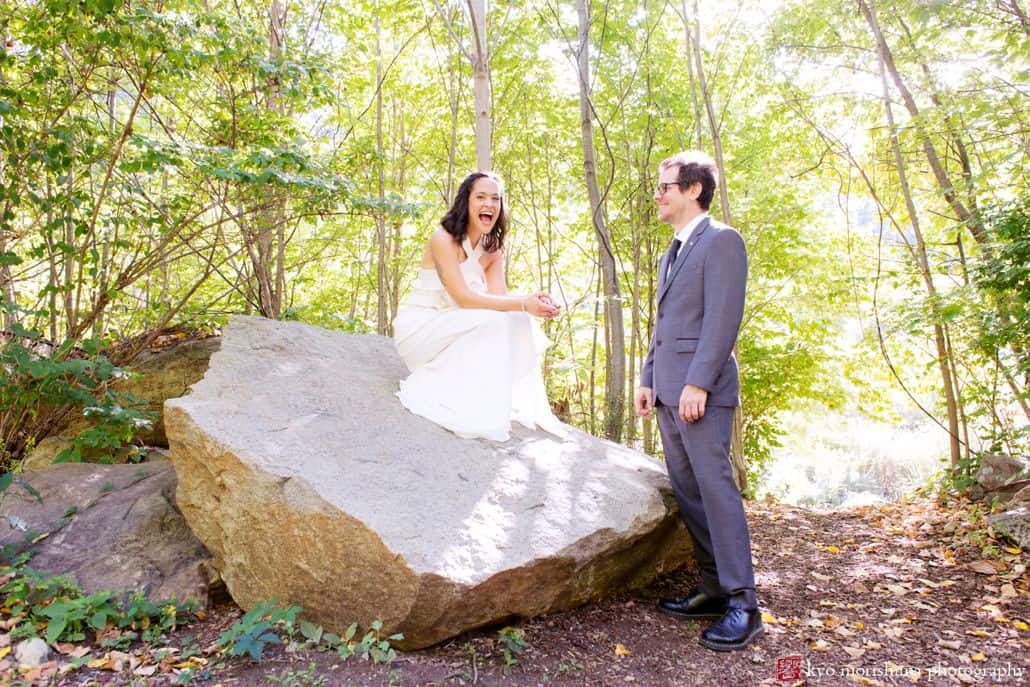 Light-hearted portrait session of a bride and groom in an outdoor location, sourounded by trees: the bride is on top of a big rock laughing and looking at the photographer and the groom is smiling looking at the bride, photographed by wedding photographer in central NJ Kyo Morishima.