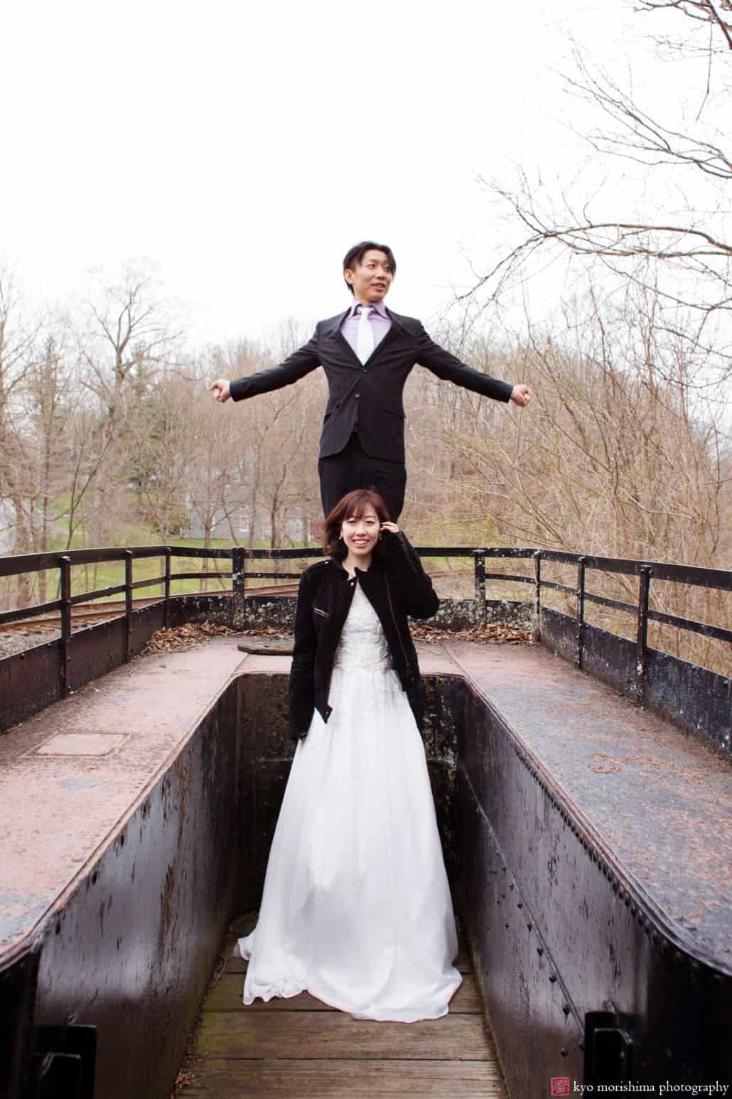 Light-hearted portrait session of a couple on an outdoor location, the groom is standing in a bench behind the bride and he's spreading his hands, while the bride is smiling wearing her wedding dress and a black jacket, photographed by wedding photographer in central NJ Kyo Morishima.