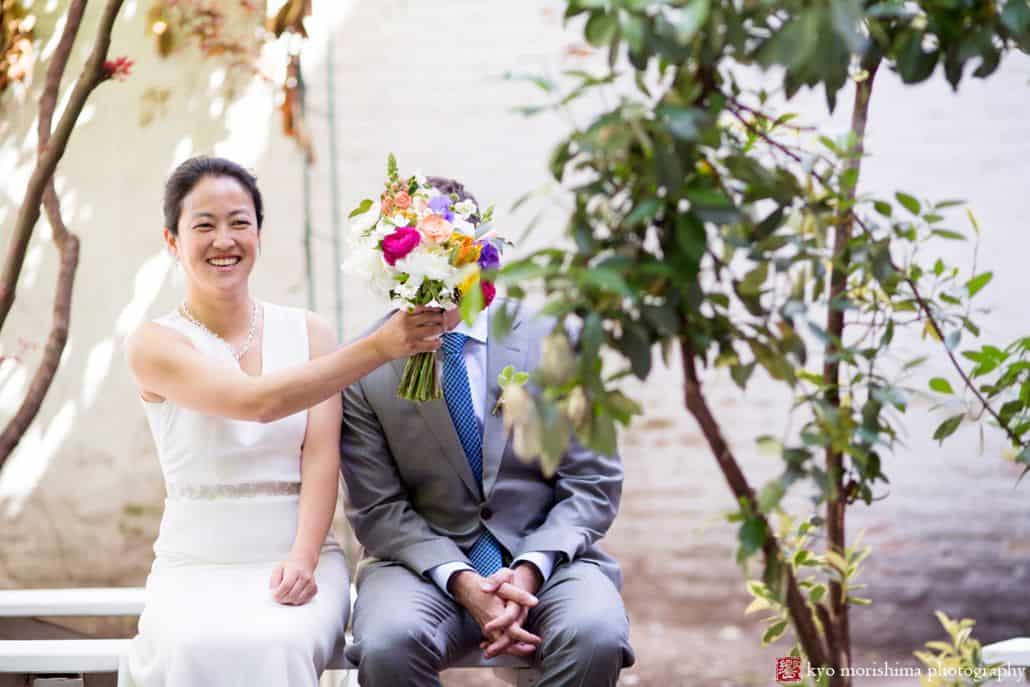 Light-hearted portrait session of a couple sitting on an outdoor bench, the bride is smiling, holding up her bouquet of flowers, covering the groom's face, photographed by wedding photographer in central NJ Kyo Morishima.