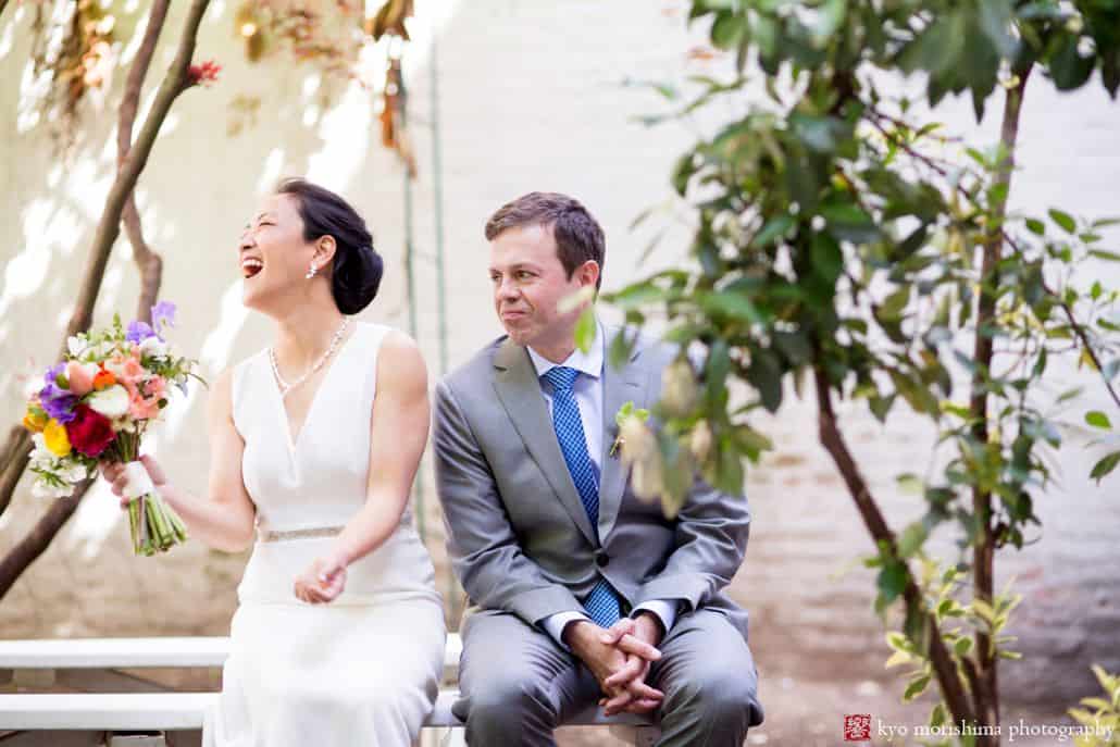 Light-hearted portrait session of a couple sitting on an outdoor bench, the bride is laughing as she holds her bouquet of flowers, and the groom is looking at the bride with a sad face, photographed by wedding photographer in central NJ Kyo Morishima.