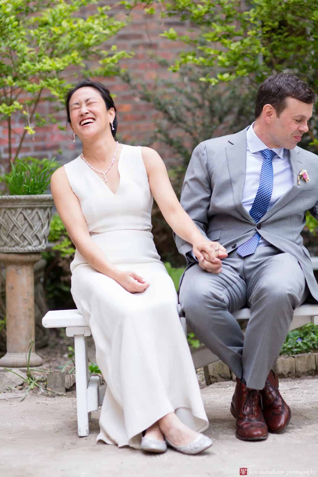 Light-hearted portrait session of a couple sitting on an outdoor bench, the bride is laughing and closing her eyes while holding the goom's hand and the groom is smiling looking at something on his left side, photographed by wedding photographer in central NJ Kyo Morishima.