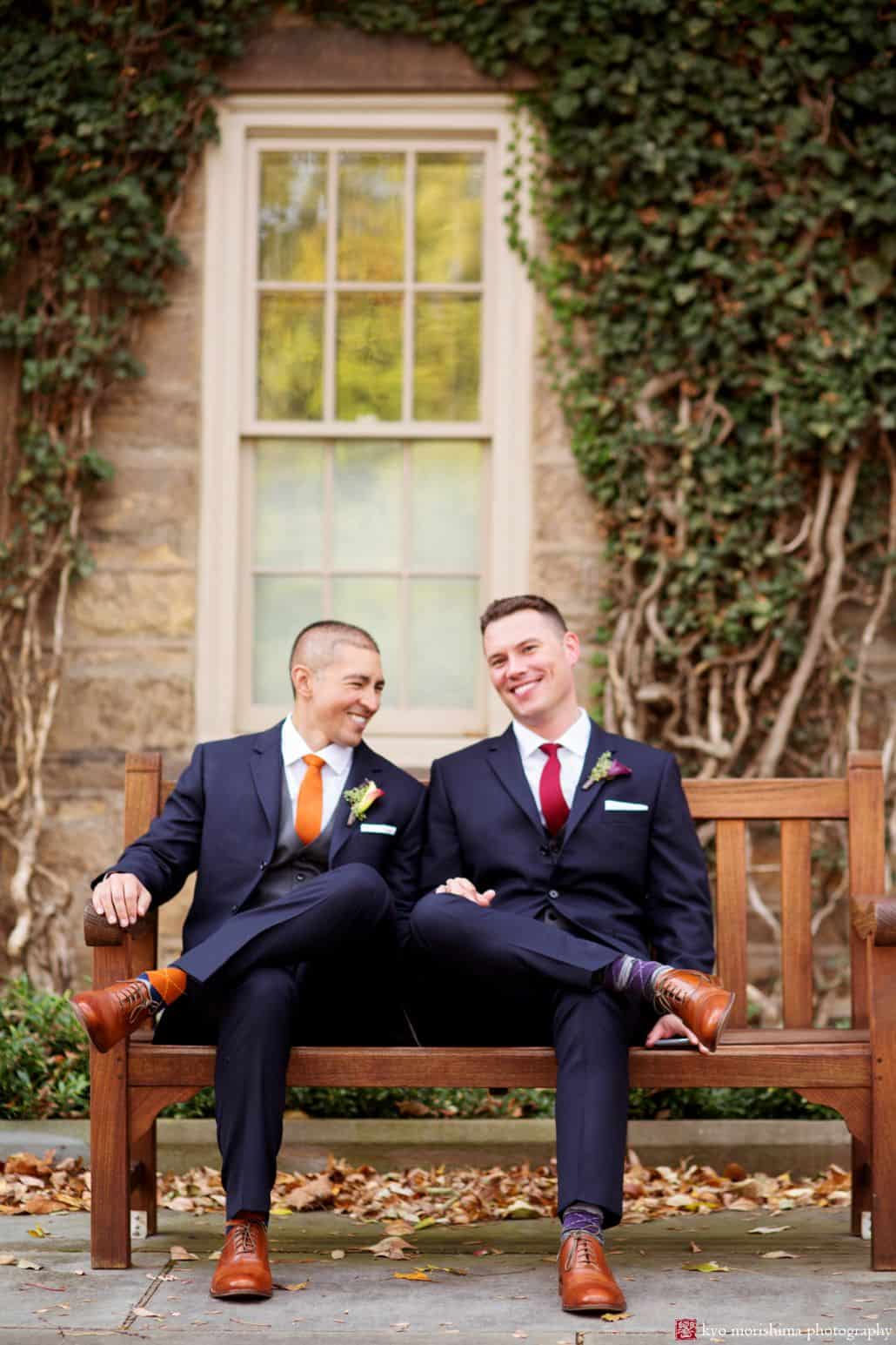 Light-hearted portrait session of two happy men sitting on a brown bench, wearing a navy blue suit and brown shoes, and they have a wall with window and vines background, photographed by wedding photographer in central NJ Kyo Morishima.