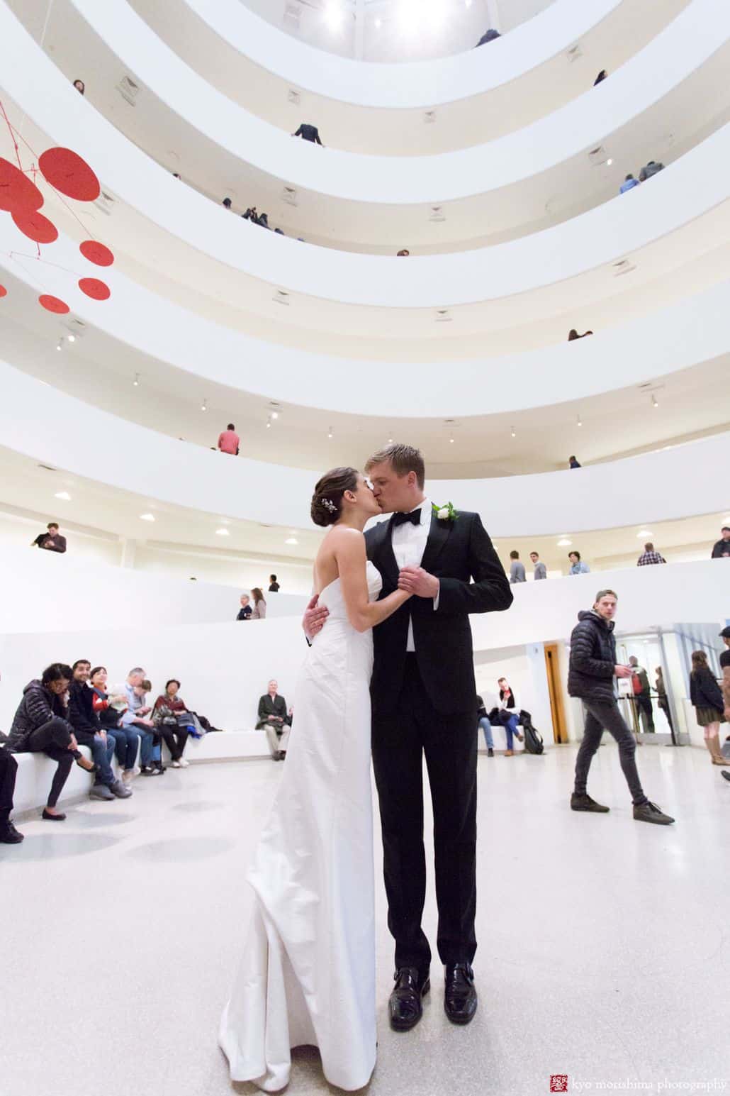 Light-hearted portrait session of a groom and bride kissing and standing in the middle of a white building's ground floor, and they are surrounded by people, photographed by wedding photographer in central NJ Kyo Morishima.