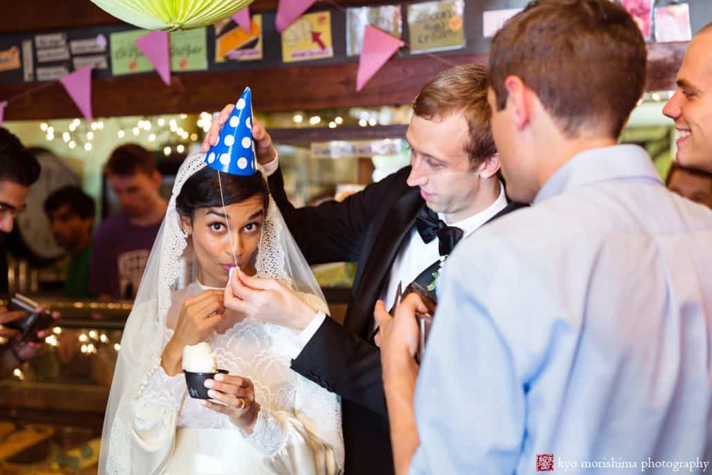 Light-hearted portrait session of the groom and bride inside a restaurant, the bride is holding a small bowl of food and the groom is putting a blue with white polka dots birthday hat to the bride's head, the guests are looking at the couple, photographed by wedding photographer in central NJ Kyo Morishima.