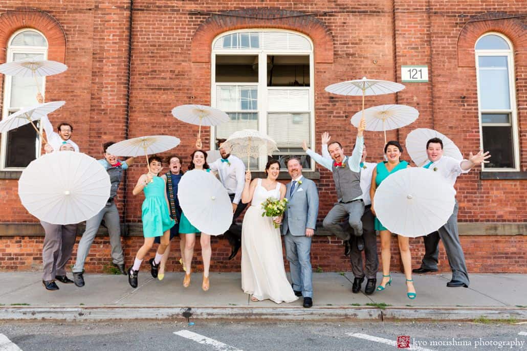Light-hearted portrait session of the happy groom and bride with their jolly guest jumping outside a brick building's sidewalk, some of the guest are holding white umbrellas, photographed by wedding photographer in central NJ Kyo Morishima.