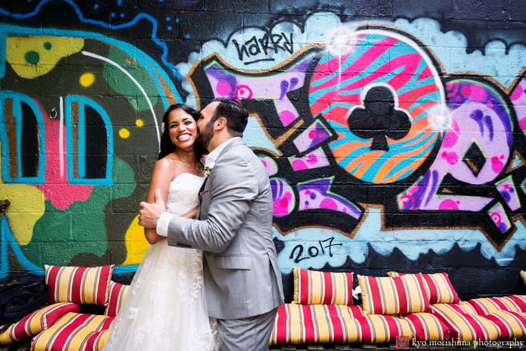 Light-hearted portrait session of a happy groom and bride, the bride is all smile groom as the groom holds her arm and kisses her on the cheeks, behind them is a wall full of graffiti and it's photographed by wedding photographer in central NJ Kyo Morishima.