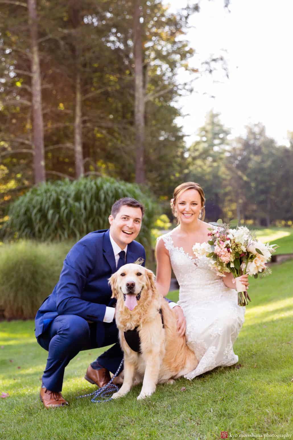Light-hearted portrait session of a happy groom and bride with their pet dog, a Labrador, the bride holding her bouquet of flowers and the groom bent down to be closer to their dog, behind them are trees and plants, photograph by a wedding photographer in central NJ Kyo Morishima.