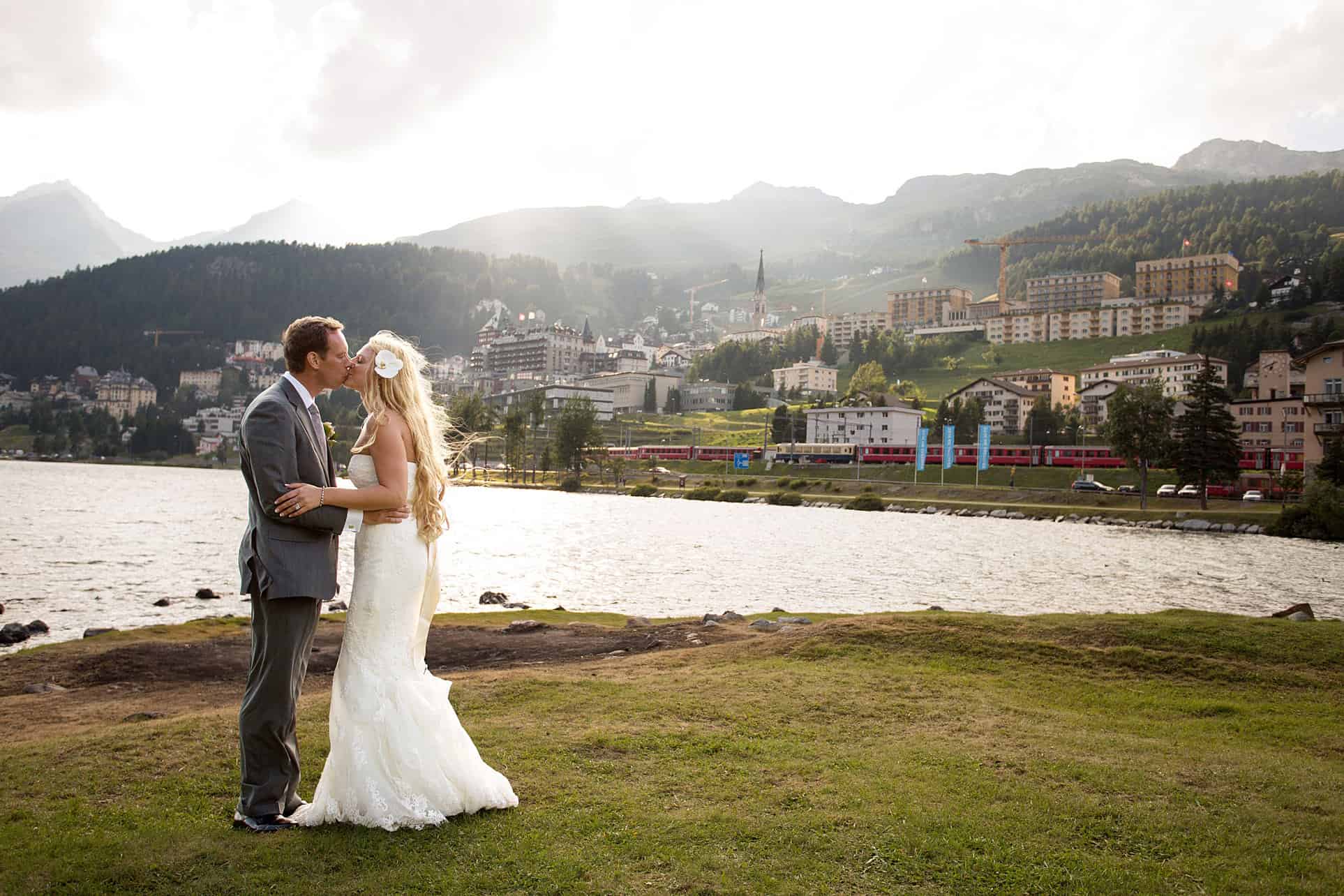 Destination wedding photographer Europe: couple kisses next to Lake St. Moritz in Switzerland. Bride wears gardenia flower in her hair and Pronovias wedding gown.