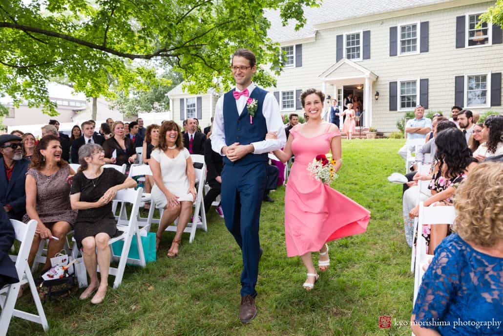 Bridesmaid and groomsman walking down the aisle wearing a pink dress and pink bow tie outside at a summer daytime wedding at the Chauncey Center.