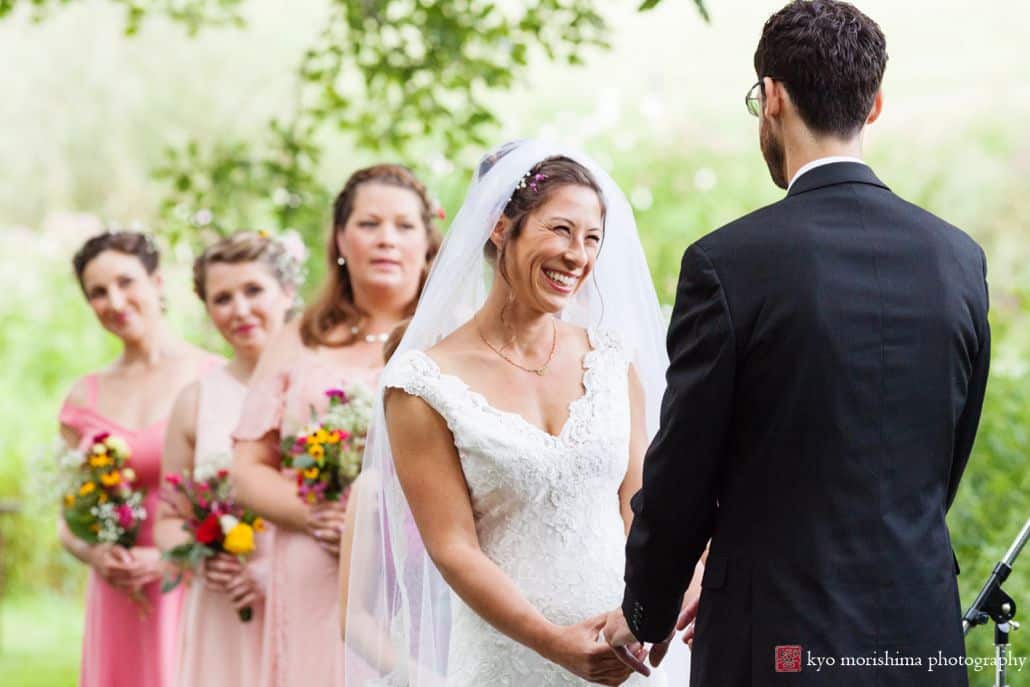 Bride smiles and holds hands with groom during outdoor summer ceremony at Chauncey Center in Princeton, with bridesmaids wearing various shades of pink looking on.