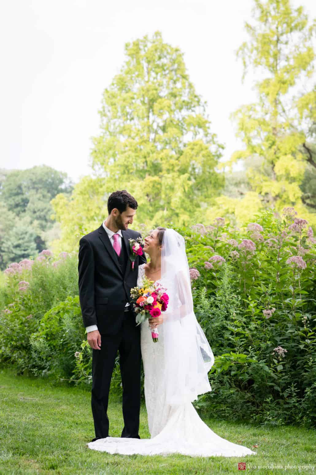 Bride and groom smile at each other during relaxed wedding portrait session at Princeton's Chauncey Center with tall flower bushes in the background.