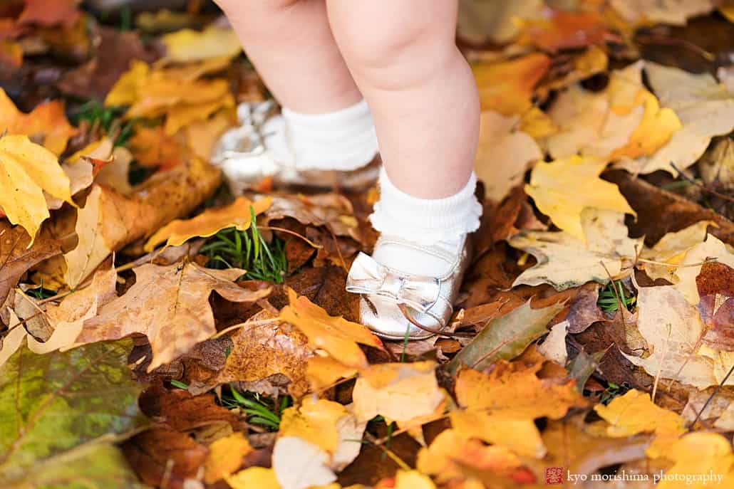 Pudgy baby legs with bowtie golden shoes standing on fall leaves: fun and candid family portrait photographer in Princeton
