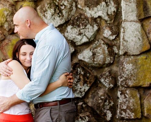Duke Farms engagement photograph: couple hugs with moss-covered stone wall in background; woman wears lipstick-red skirt