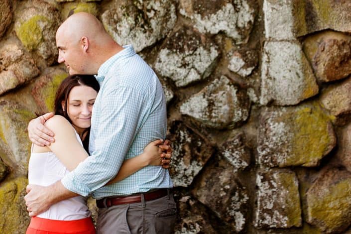 Duke Farms engagement photograph: couple hugs with moss-covered stone wall in background; woman wears lipstick-red skirt