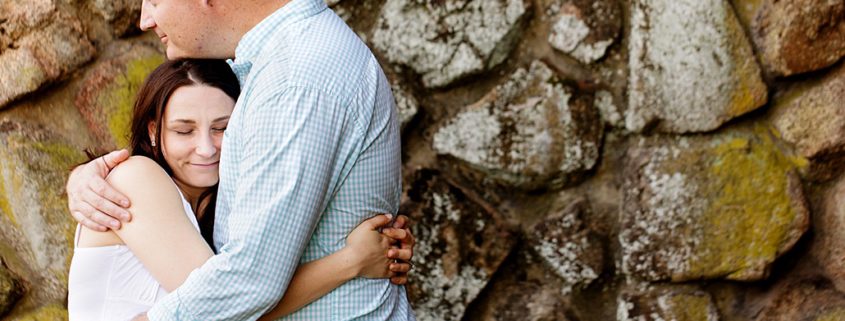 Duke Farms engagement photograph: couple hugs with moss-covered stone wall in background; woman wears lipstick-red skirt