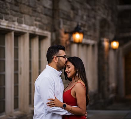 Princeton University Campus Firestone Library NJ candid documentary engagement portrait session fall autumn a couple red dress European brick building wooded door street lights hold each other hug kiss on her fore head smile
