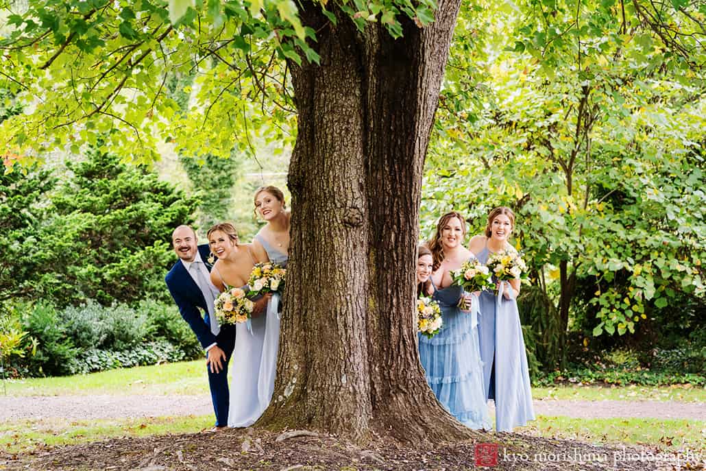 bridesmaids in blue dress and suit having fun holding bouquets getting together smiling laughing behind a huge big tree hiding at Fall The Inn at Fernbrook Farm Chesterfield NJ Wedding